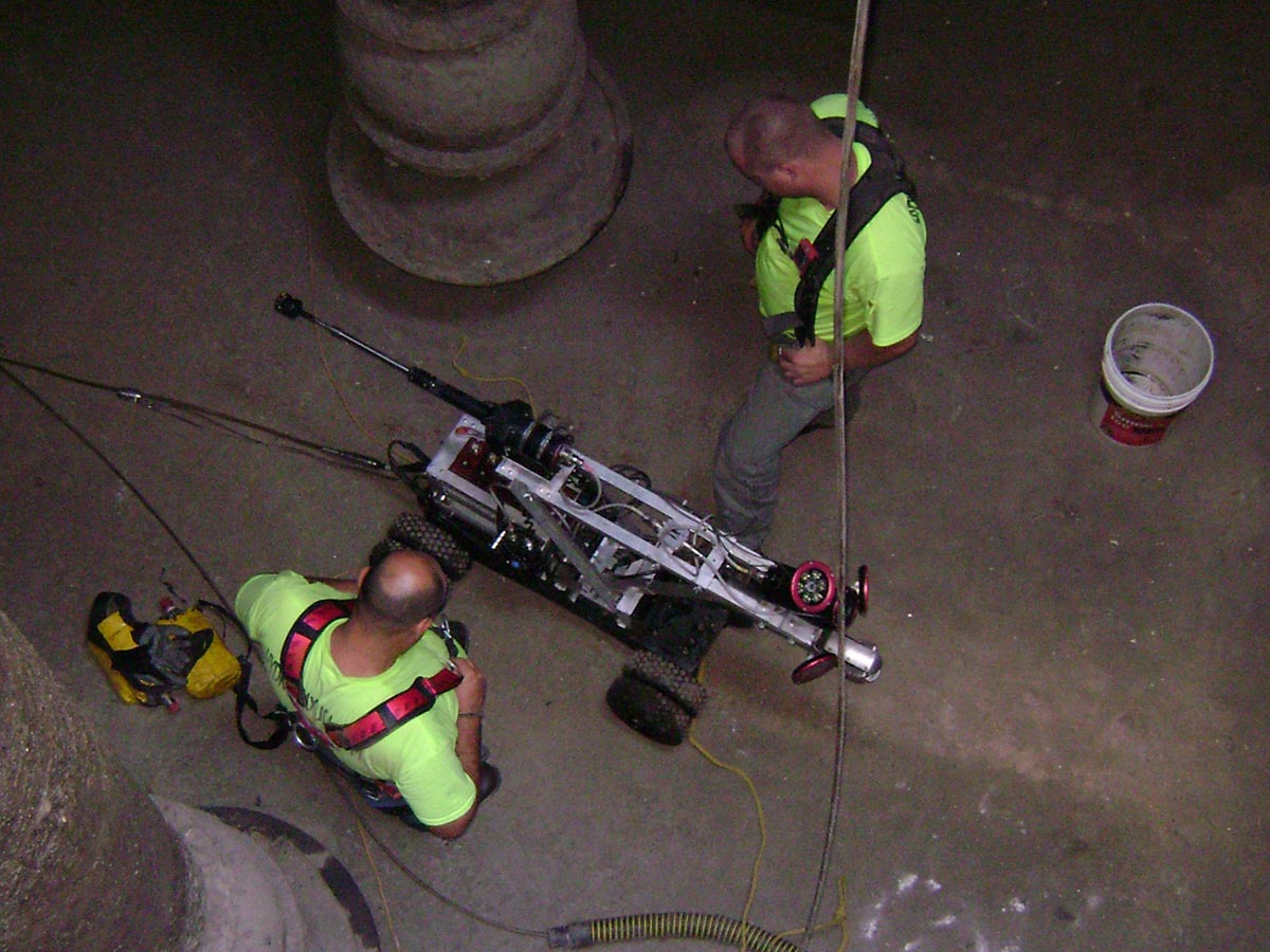 Two Hydromax workers inside a sewer system, examining a wheeled sensor drone during an inspection